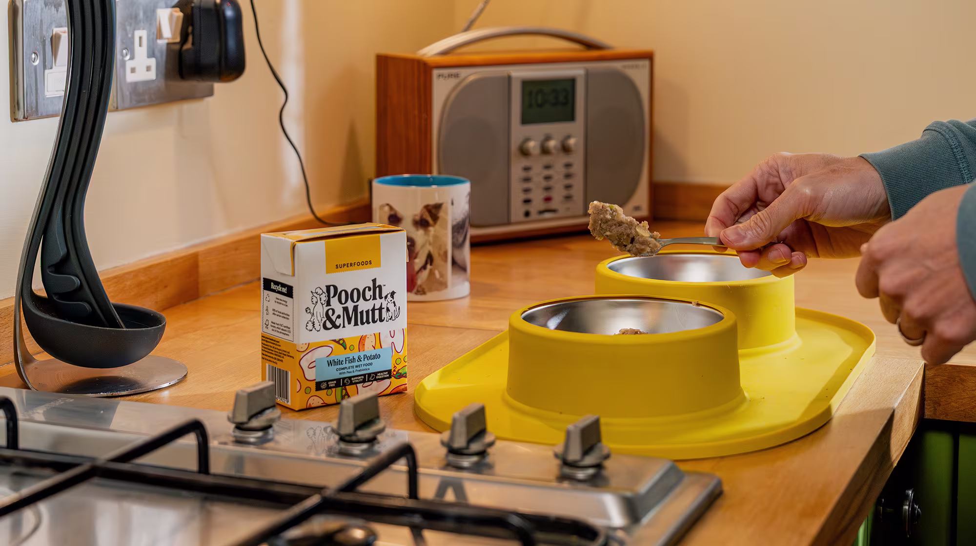 A dog owner spooning dog food into a dog bowl on the kitchen counter, with a pack of Pooch & Mutt's White Fish & Potato Wet Food next to it.