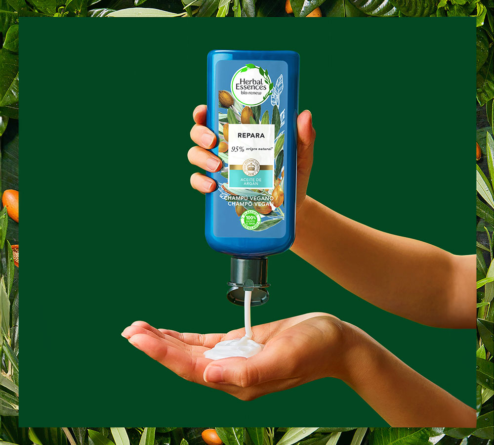 Una mano exprime un tubo de Herbal Essences Champú Reparador, dispensando producto blanco en otra mano.