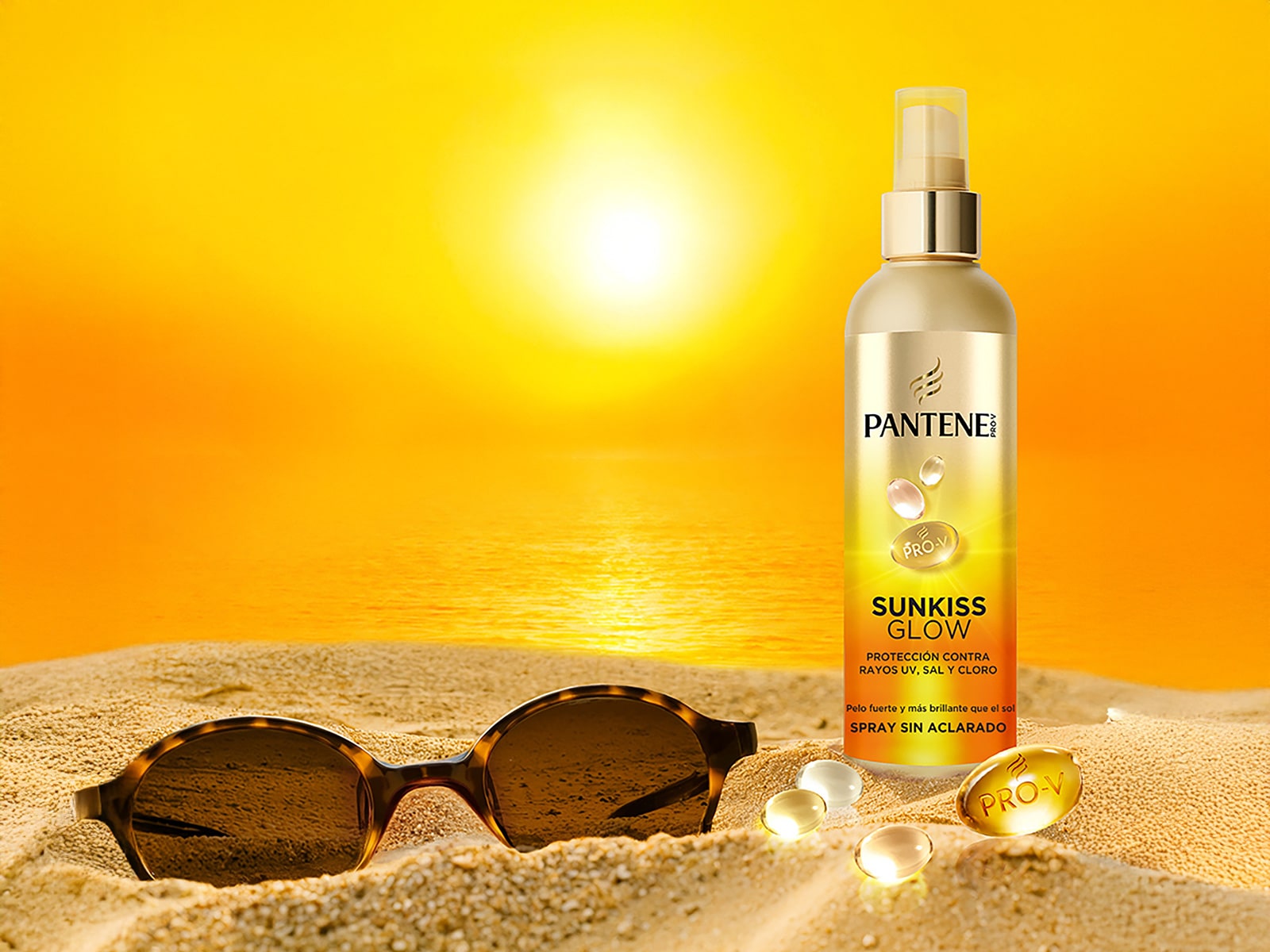 Spray Pantene Sunkiss Glow sobre arena con gafas y gotas doradas, atardecer con sol reflejado en agua.