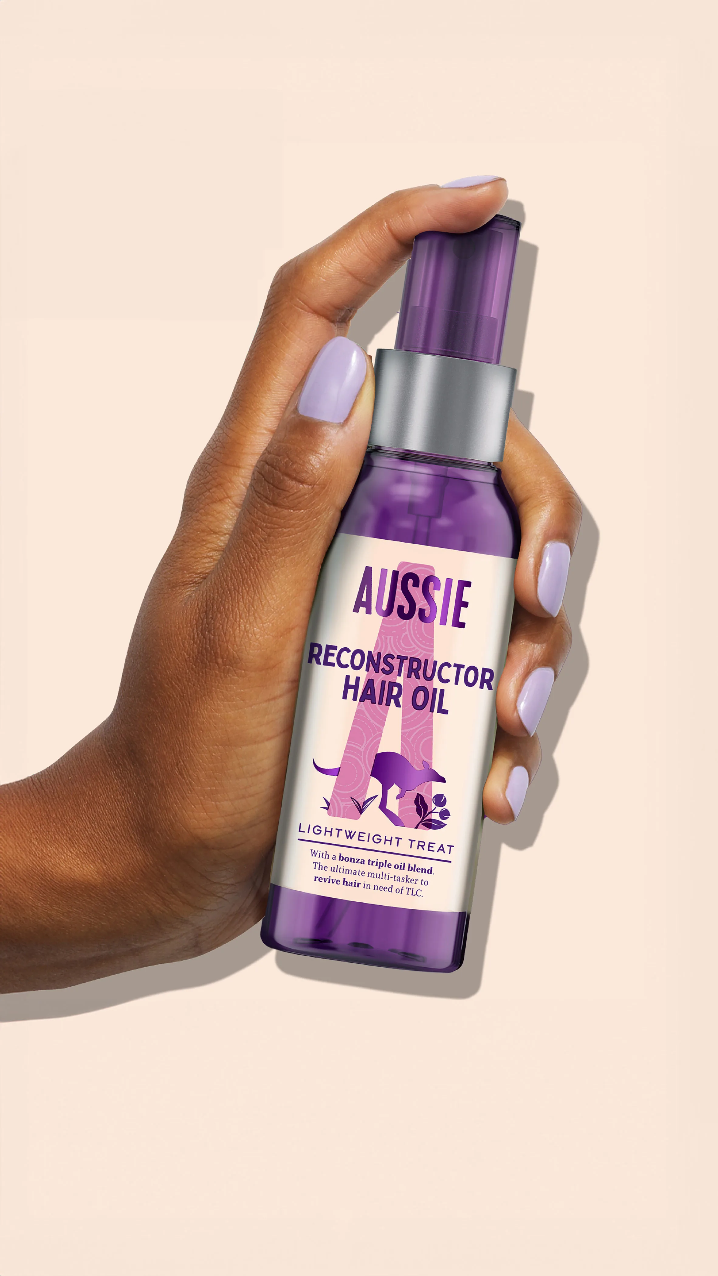 Mano con uñas pintadas sostiene botella morada de Aussie Aceite Reconstructor sobre fondo beige claro.