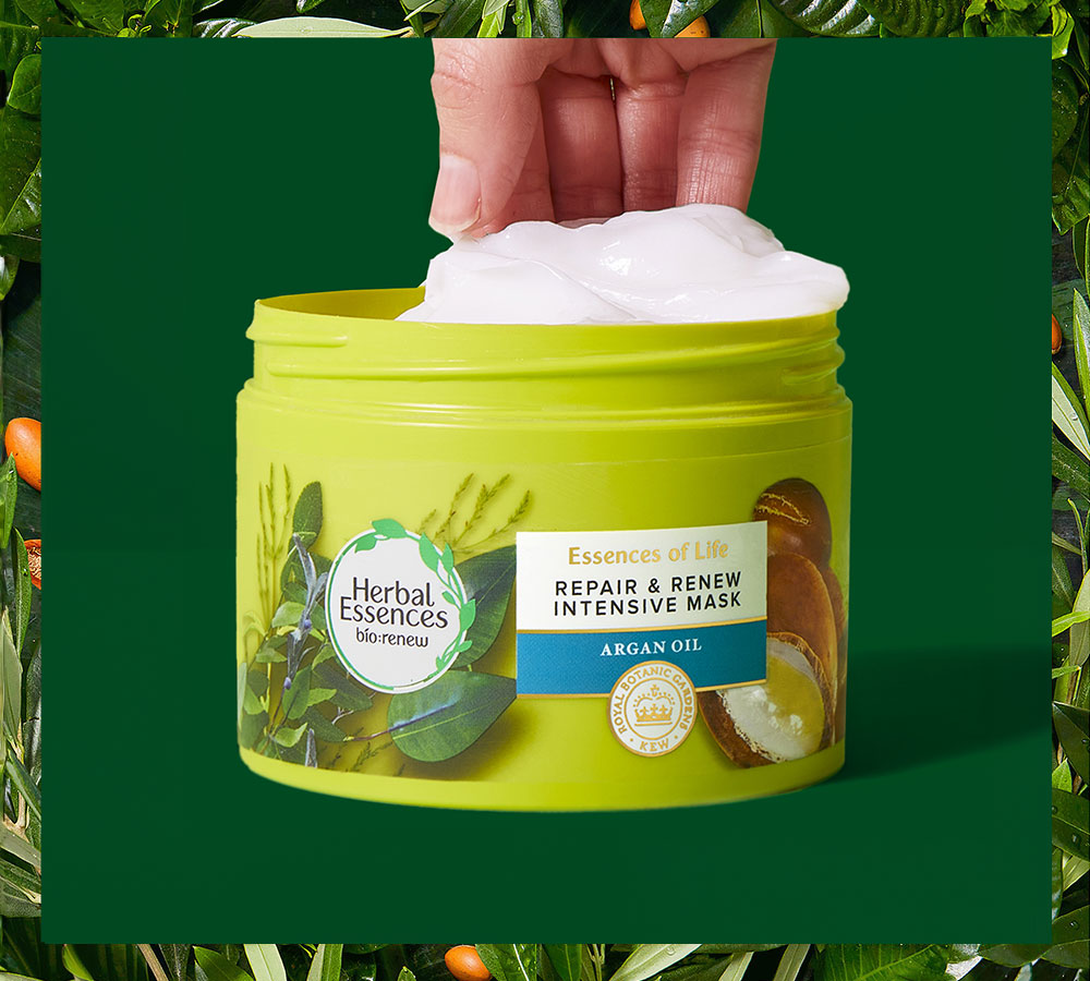 Una mano saca producto blanco de un tarro verde de Mascarilla Herbal Essences Repair & Renew, rodeado de hojas verdes.