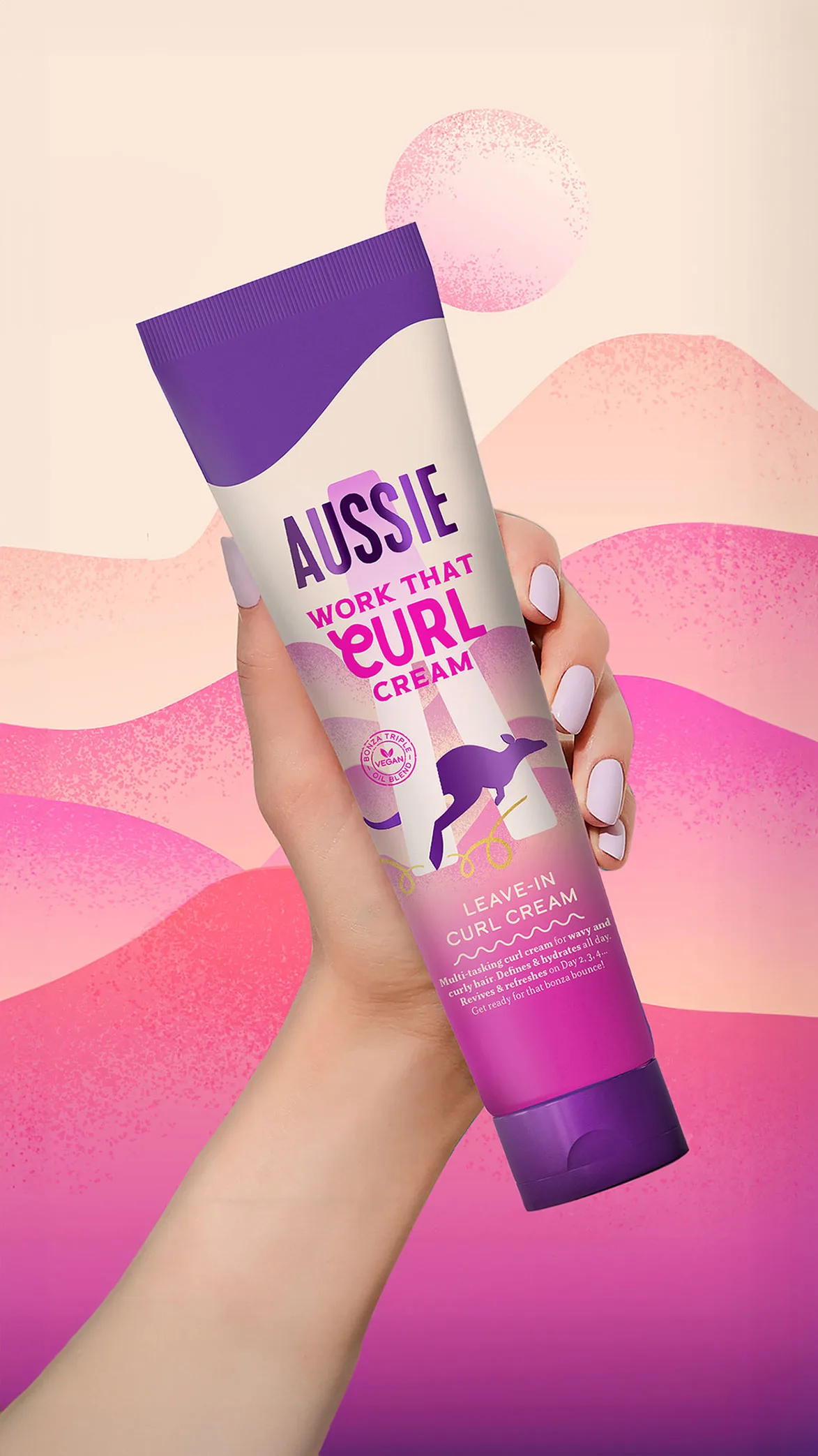 Una mano sostiene un tubo de Aussie Work That Curl Crema con etiqueta blanca, canguro morado, sobre fondo rosa y morado ondulado.