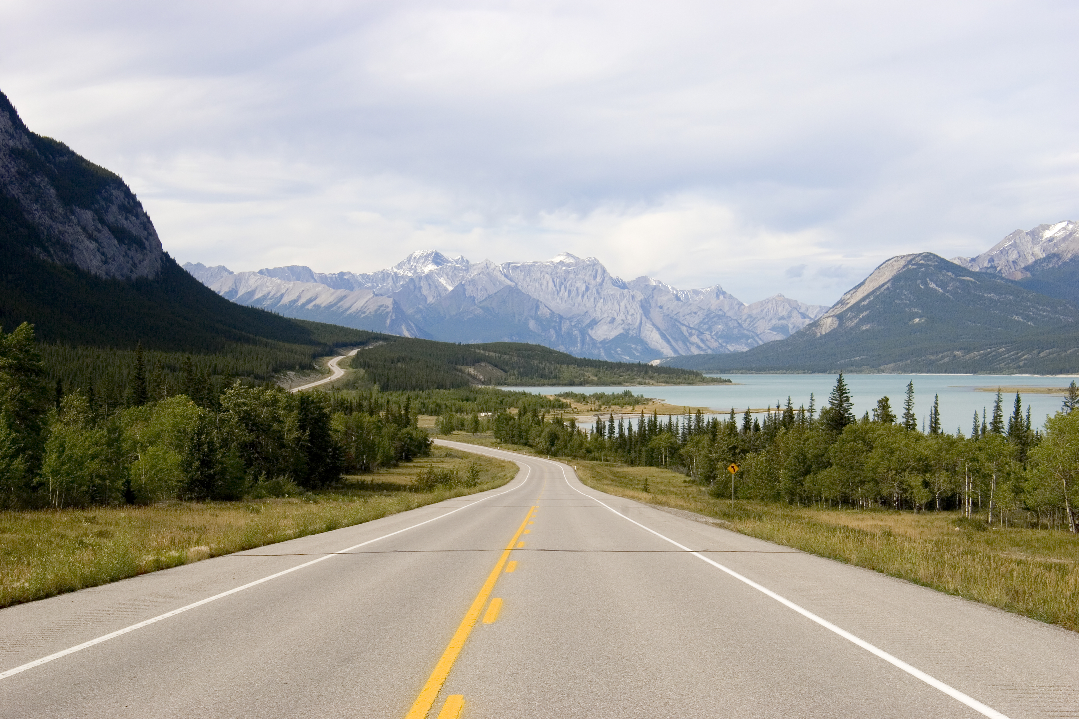 A two‑lane road winds through a vast natural landscape in Western Canada, surrounded by dense forests, towering mountains, and a calm lake. The scenery reflects the iconic environments of Alberta and British Columbia, where panoramic highways meet expansive wilderness. Beneath a bright, lightly clouded sky, this image conveys summer driving, road travel, and the dramatic beauty of Canada’s mountain regions.
