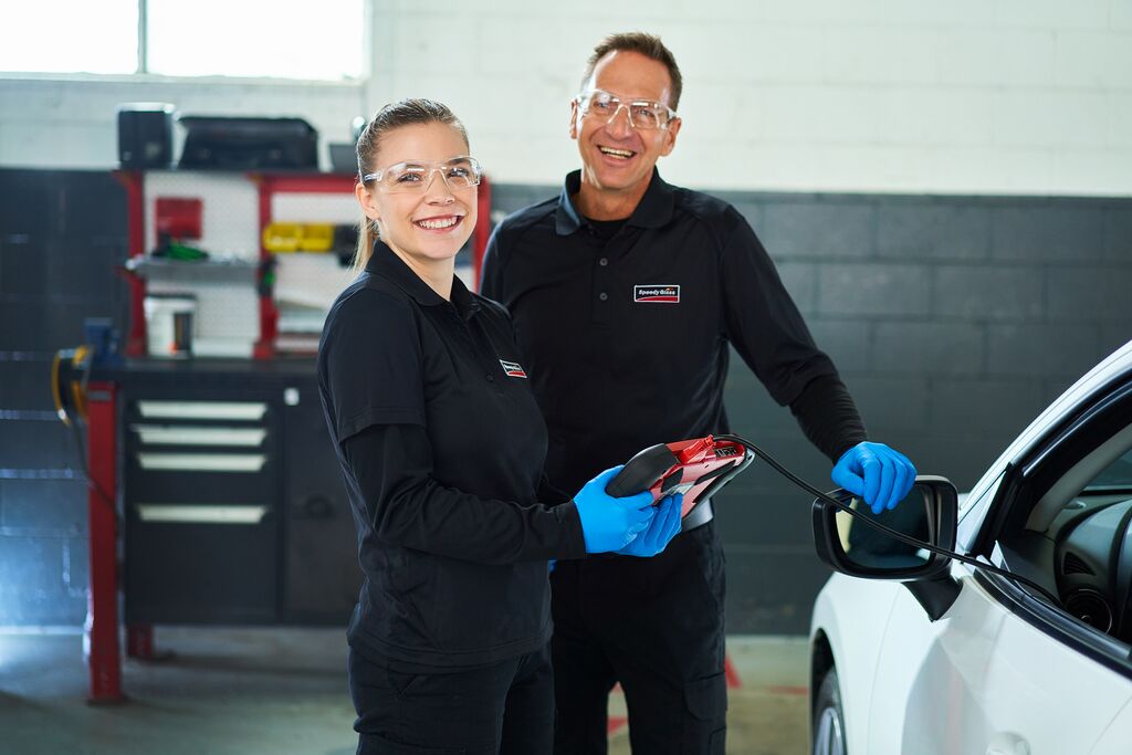 Two smiling technicians in a garage, holding tools and working on a vehicle