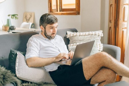 A man sitting on a couch while working from home with a laptop in his lap.