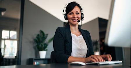 Help desk professional wearing a headset, smiling and typing at a desk in a modern office.