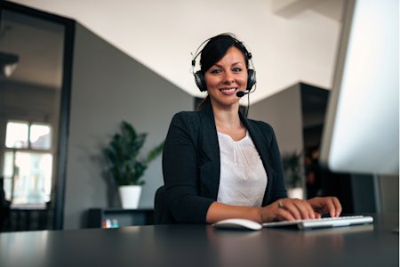 Help desk professional wearing a headset, smiling and typing at a desk in a modern office.