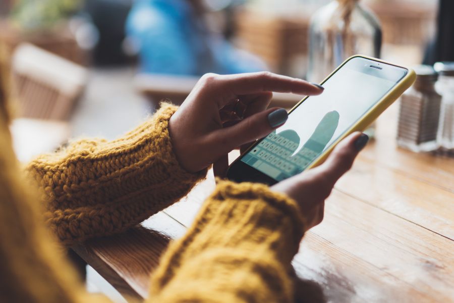 A person wearing a yellow sweater sits at a wooden table, using a smartphone with both hands. The phone screen displays a messaging app keyboard, and the background is softly blurred.