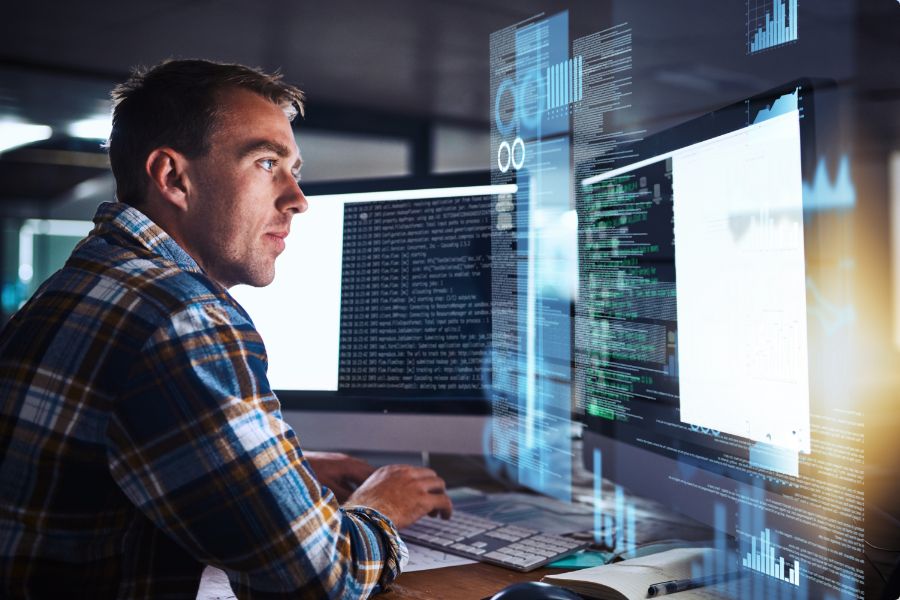 A man in a plaid shirt sits at a desk, focusing on computer screens displaying lines of code and data visualizations in a modern office setting.