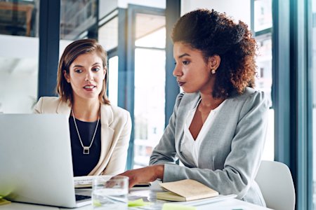 Two businesswomen collaborating on a laptop during an IT risk assessment meeting in a modern office.