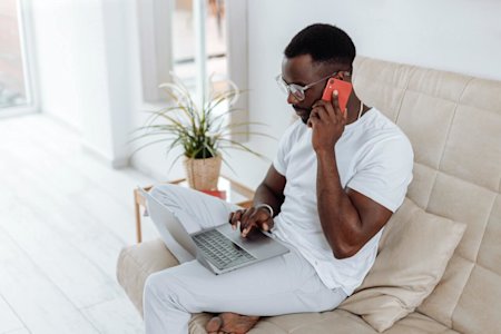A man sitting on a couch with a laptop and talking on a smartphone while working remotely.