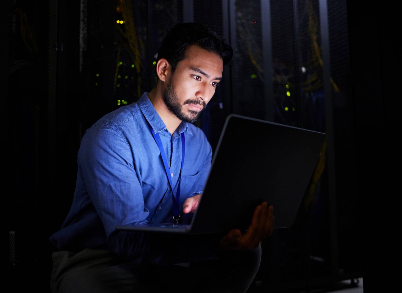 A man in a blue shirt sits in a dimly lit server room, focused on working with a laptop, with computer servers and cables visible in the background.
