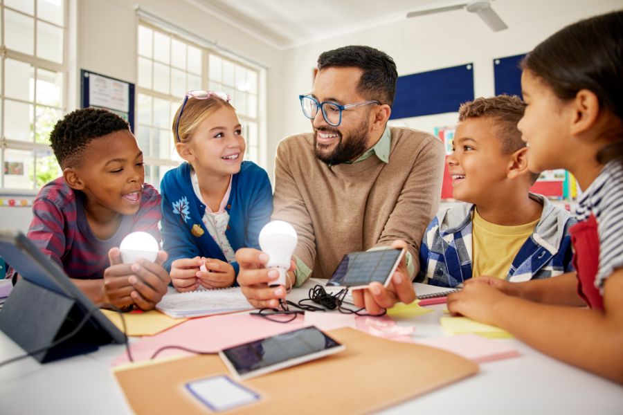 A teacher sits at a table with four smiling students holding light bulbs, engaging in a science experiment with wires, a tablet, and notebooks in a bright classroom.