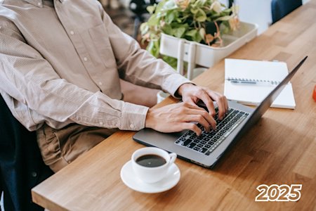 Person working remotely on a laptop at a wooden table with a coffee cup and notebook, symbolizing remote work trends in 2025.