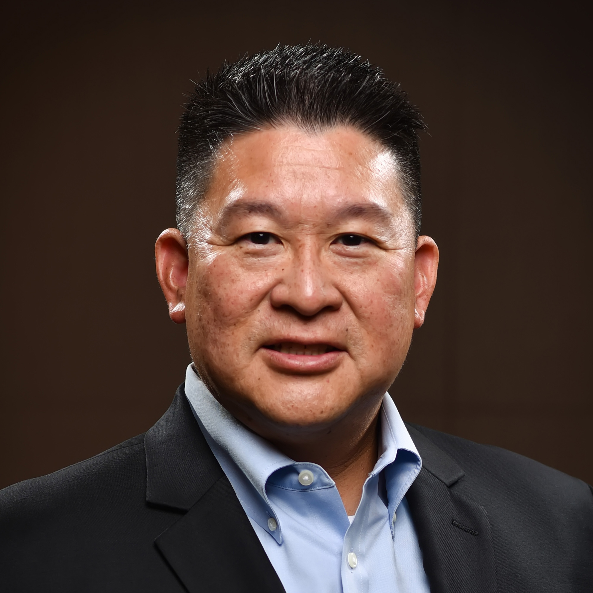 A man with short black hair wearing a dark suit jacket and light blue collared shirt, smiling slightly, posed against a dark, plain background.