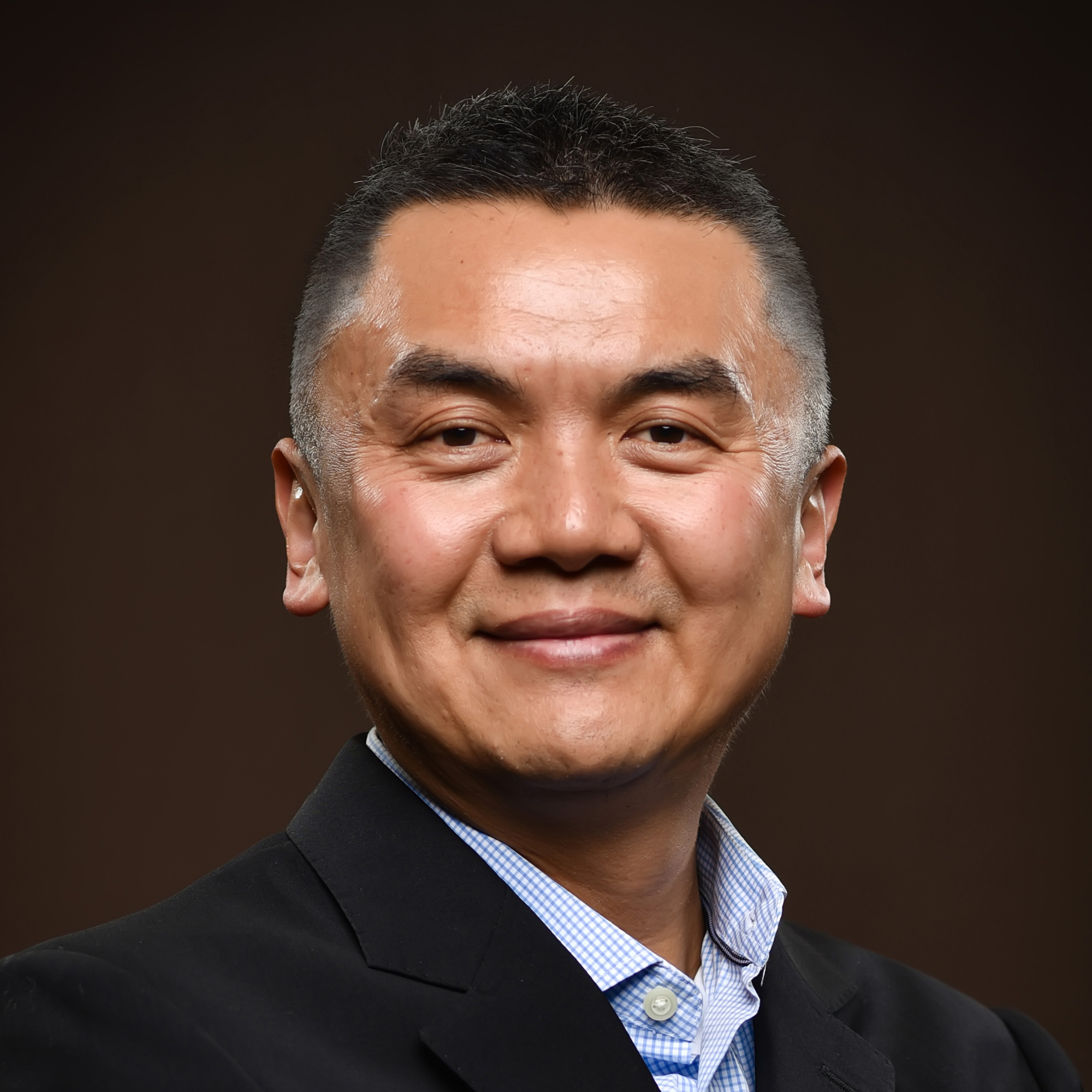 A man with short black hair and a slight smile, wearing a black blazer over a light blue shirt, poses against a dark brown background.