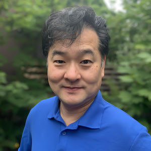 A man with short, dark hair wearing a blue collared shirt smiles at the camera while standing outdoors in front of green foliage.