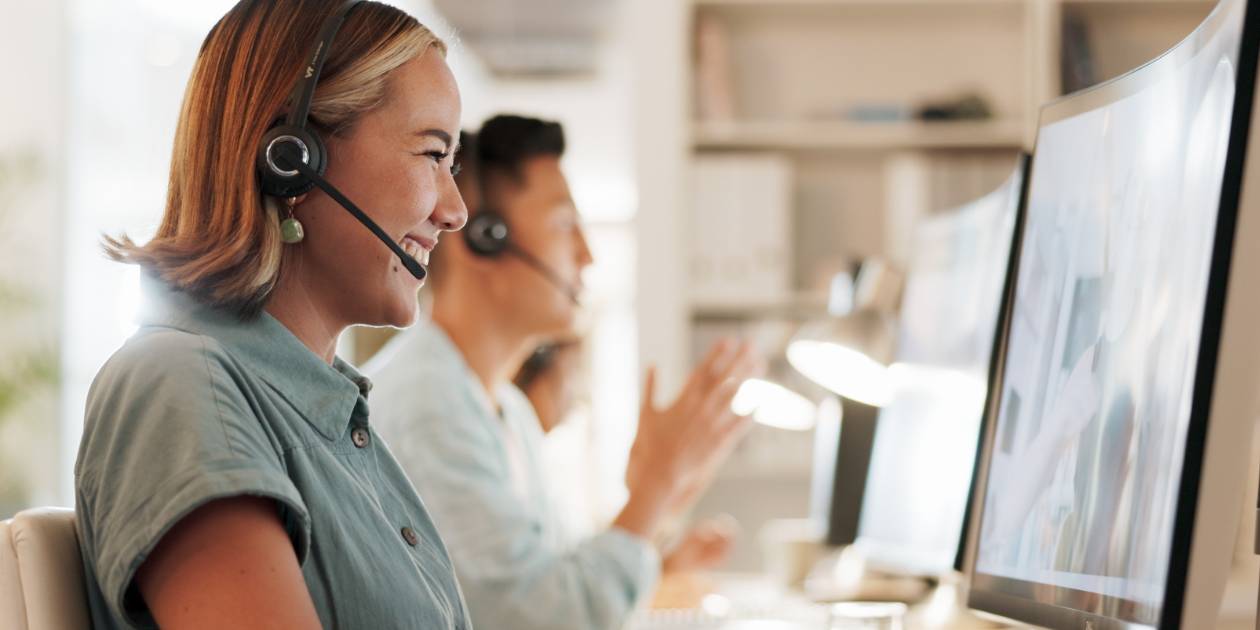Two call center employees wearing headsets sit at desks, smiling and talking while using computers in a brightly lit office environment.