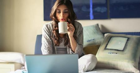 A woman sitting on her bed and drinking a coffee while using a laptop to access a remote desktop.