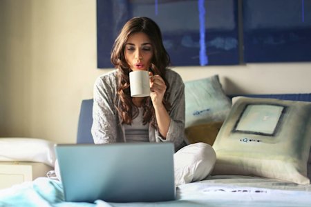 A woman sitting on her bed and drinking a coffee while using a laptop to access a remote desktop.