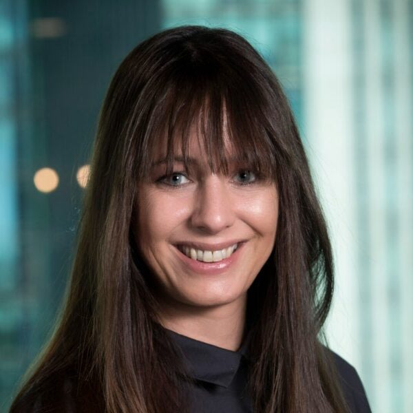 A woman with long, straight brown hair and bangs smiles at the camera. She is wearing a dark top and is posed in front of a blurred office window background.