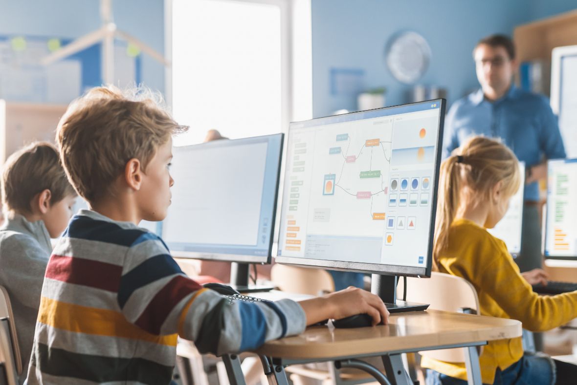 Children sit at desks in a classroom, working on computers with colorful diagrams on the screens. A teacher stands in the background, overseeing the students’ activities. The room is bright and modern.