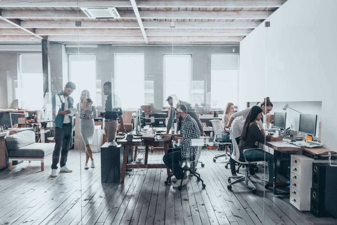 Modern open-plan office with people working at desks and computers, while some colleagues stand and talk. The space has large windows, wooden floors, and exposed ceiling beams, creating a bright, collaborative environment.