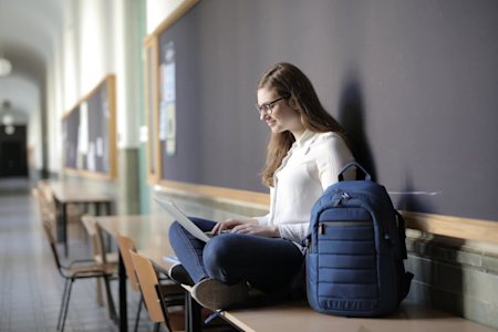 A female student sitting on a desk with her laptop in her lap and a backback next to her.