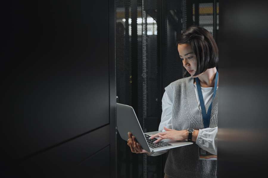 A woman stands in a server room, working on a laptop. She wears a lanyard and business attire, and is focused on the screen as she types, surrounded by tall black server racks.