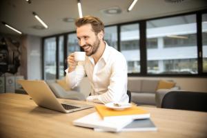 A smiling man sipping coffee while working at his laptop.