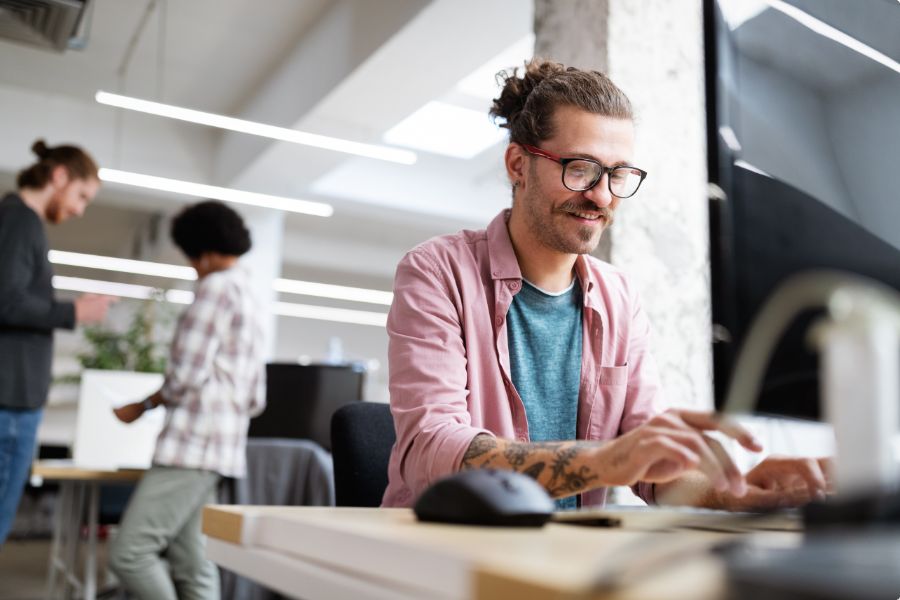 A man with glasses and a bun smiles while working on a computer at a desk in a modern office. Two other people are seen collaborating in the background.