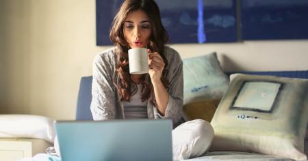 A woman sitting on her bed working on a laptop an drinking coffee.