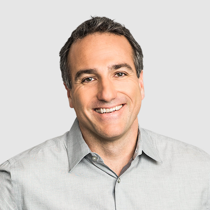A man with short dark hair and a light gray button-up shirt smiles at the camera against a plain light gray background.