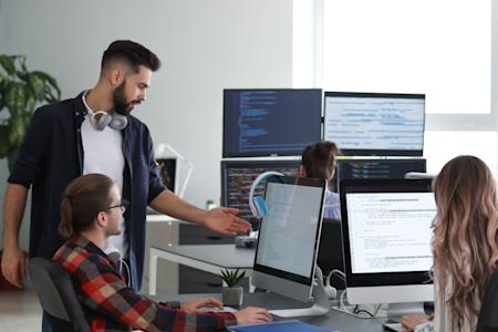 Four people work together in a modern office with multiple computer monitors displaying code; one man stands and gestures at a colleague’s screen, while others sit and focus on their tasks.