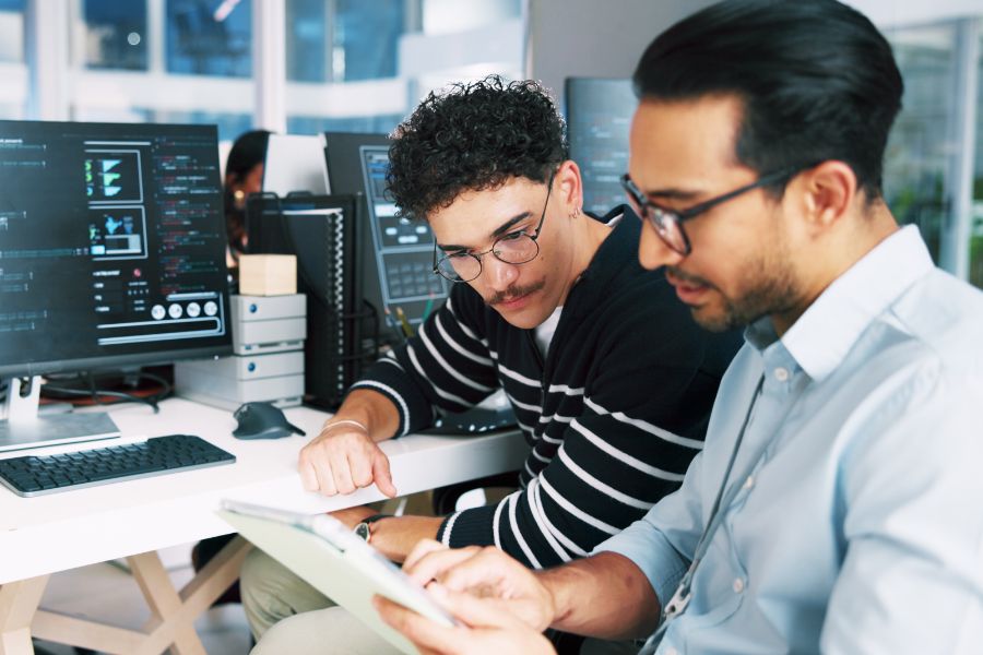 Two men working at a desk with multiple computer monitors displaying code. One man is looking at a digital tablet held by the other man, and they appear to be discussing something work-related.