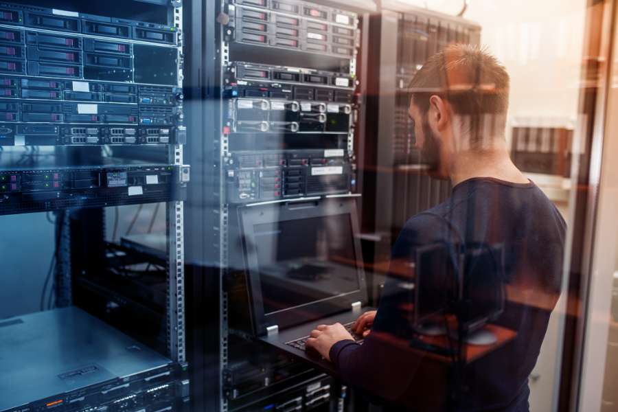 A man works on a laptop in a server room, surrounded by racks of network servers and equipment, with reflections of technology visible on the glass.
