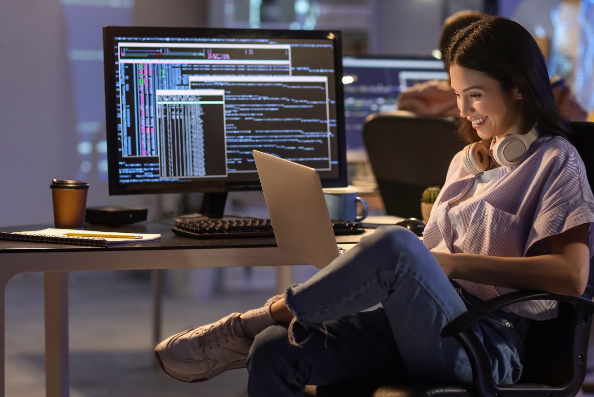 A woman sits at a desk with a laptop, smiling while working. She has headphones around her neck. Behind her are two monitors displaying code. A coffee cup and notepad are also on the desk.