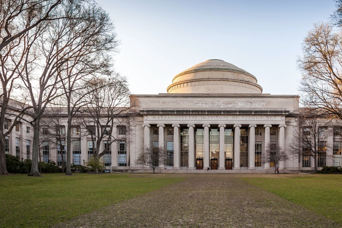 The front view of the Massachusetts Institute of Technology (MIT) building features tall columns, a large dome, and leafless trees, with a green lawn in the foreground.