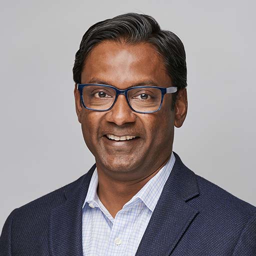 A man wearing dark blue glasses, a dark blazer, and a light checkered shirt smiles in front of a plain light gray background.