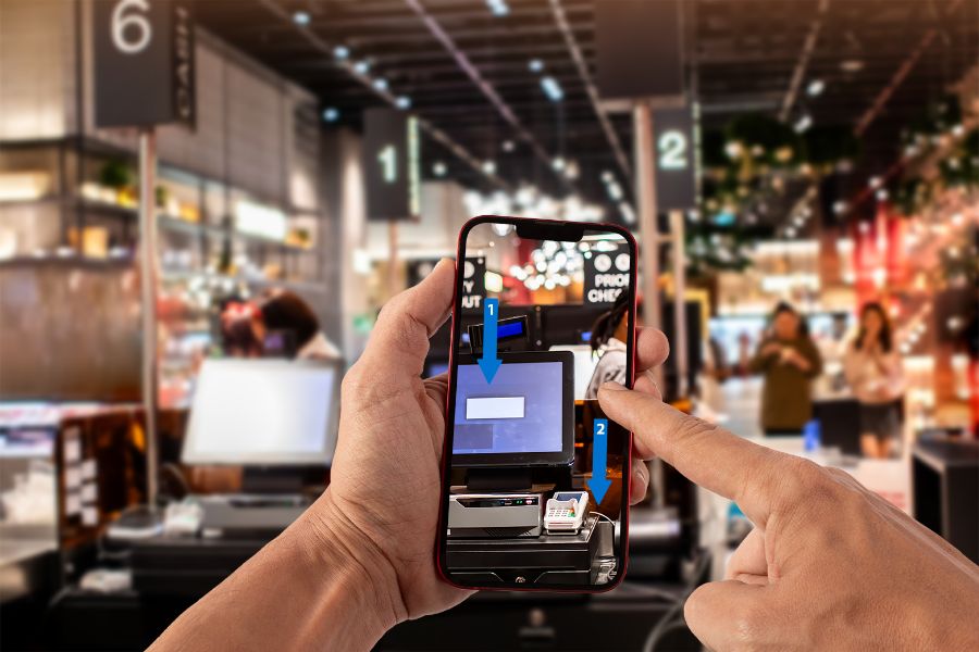 A person holds a smartphone displaying an augmented reality interface, pointing at a self-checkout machine in a brightly lit store with blurred shoppers and signage in the background.