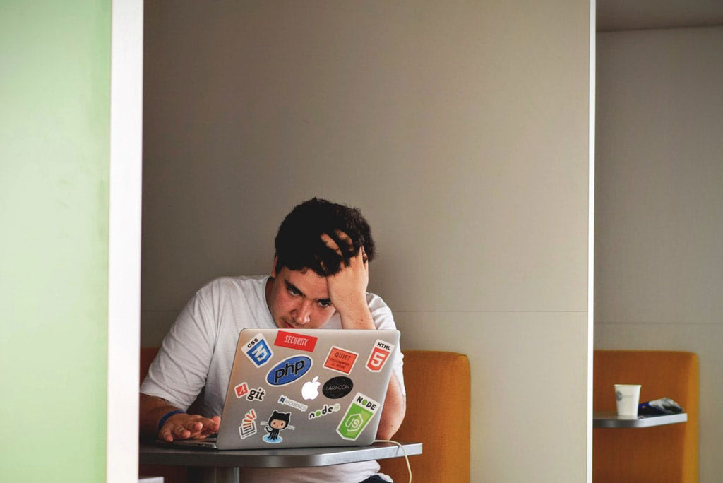 A person sits at a small table, looking stressed with their hand on their forehead, working on a laptop covered in tech and programming stickers. There is a paper cup on a nearby table.