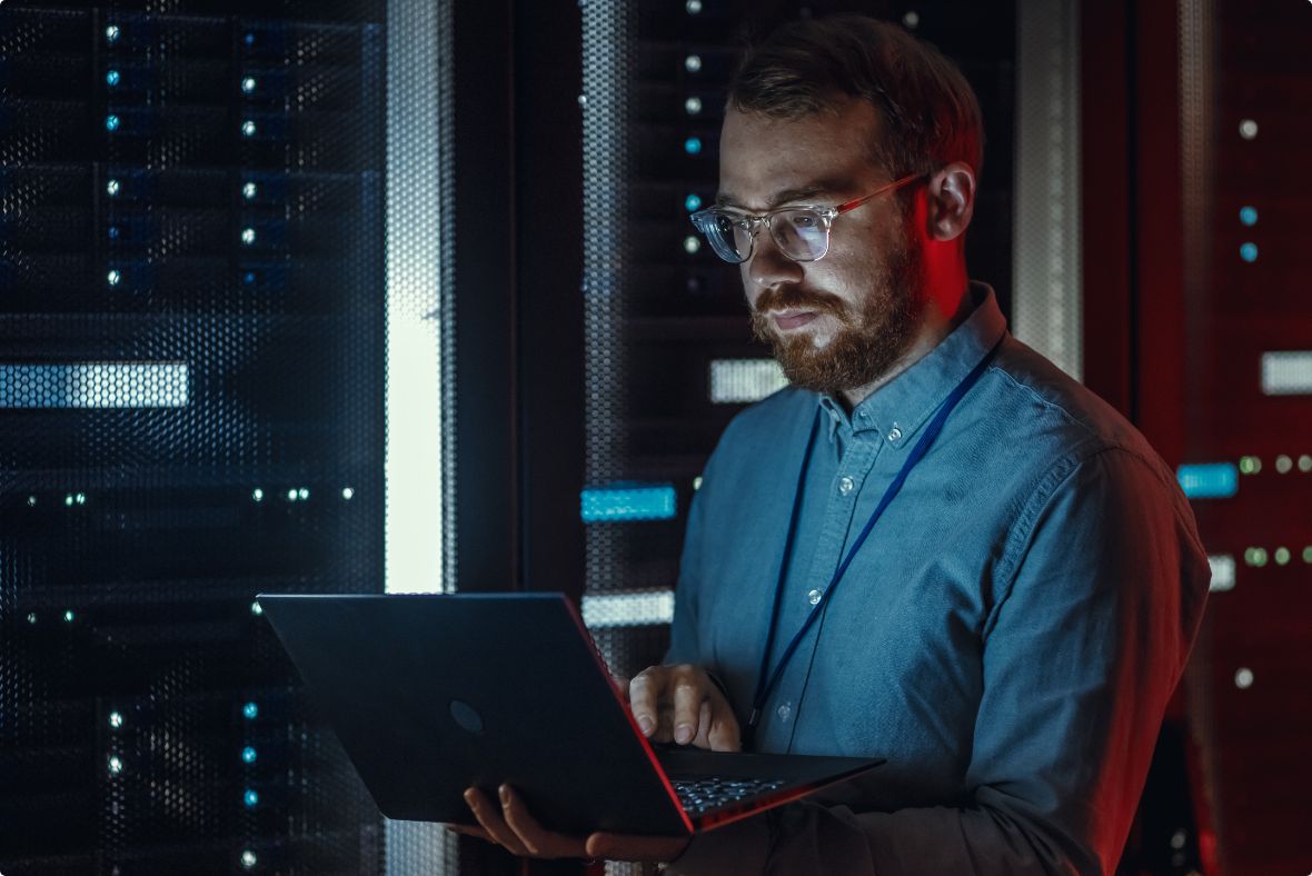A man in a server room works on a laptop, illuminated by the cool glow of rack lights while focusing intently on whatever problem he’s trying to untangle.