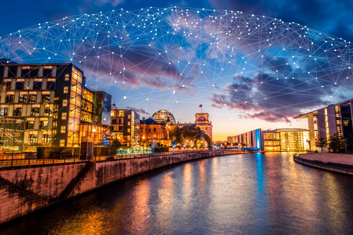 A cityscape at dusk with a river in the foreground, modern and historic buildings on both sides, and an overlay of glowing network lines and points forming a digital web in the sky above.