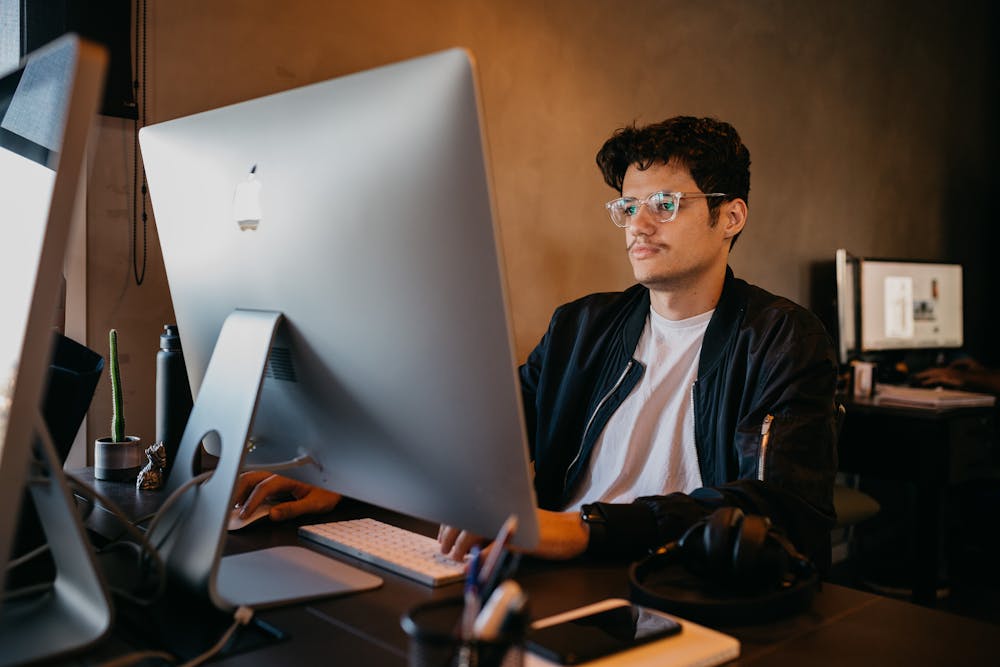 A man wearing glasses and a dark jacket works at a large desktop computer in a modern office, with a keyboard, headphones, and desk accessories visible around him. Another computer is in the background.