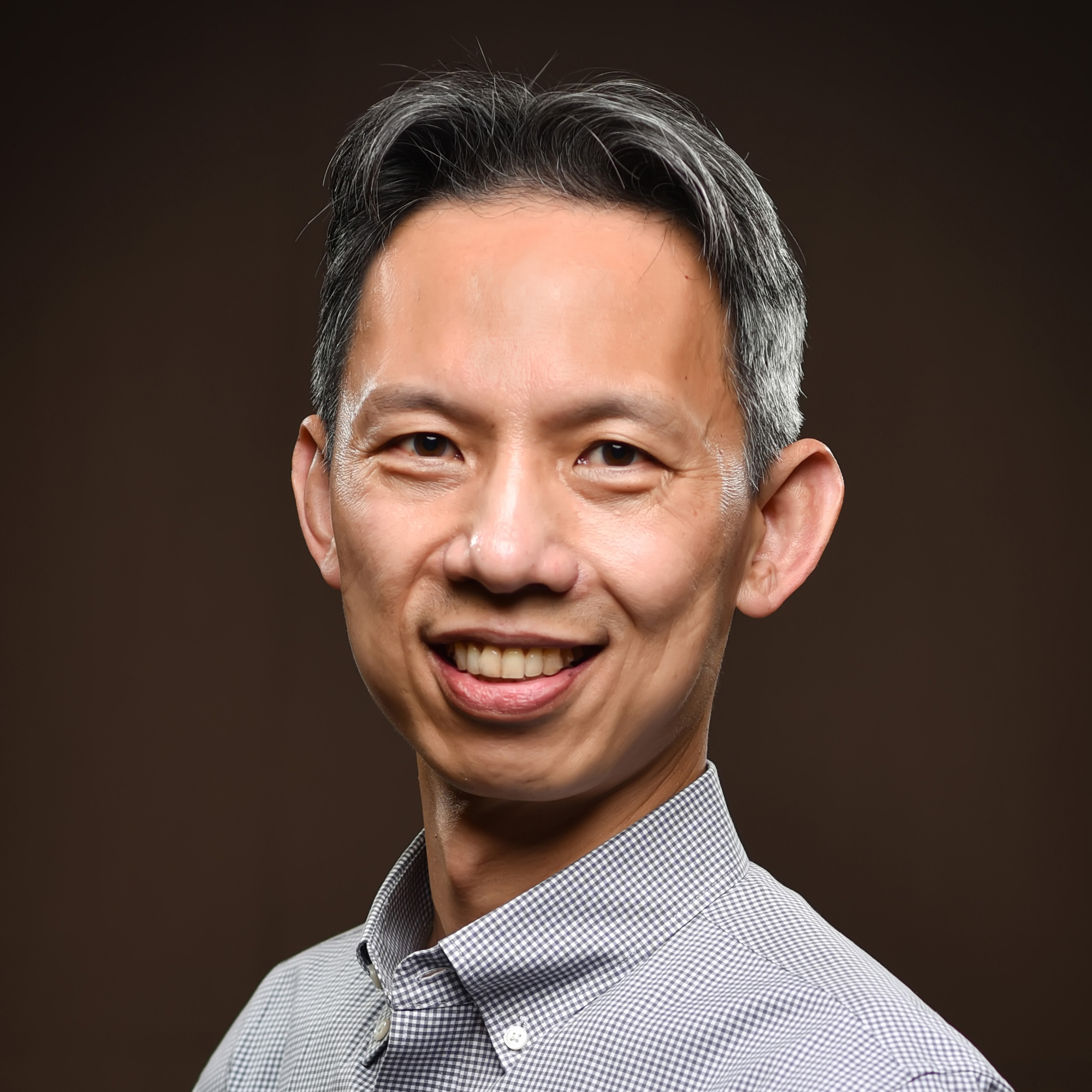 A man with short gray and black hair, wearing a light checkered shirt, smiles at the camera against a plain dark background.