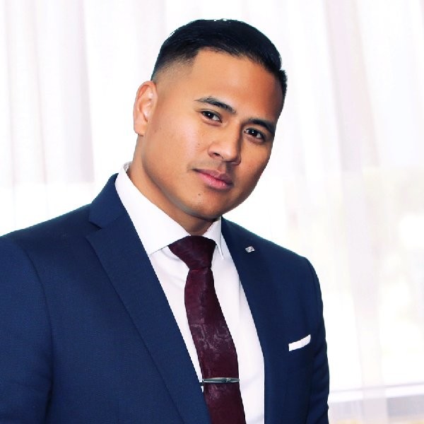 A man in a navy blue suit, white shirt, and dark red tie stands in front of sheer white curtains, looking at the camera with a slight smile.