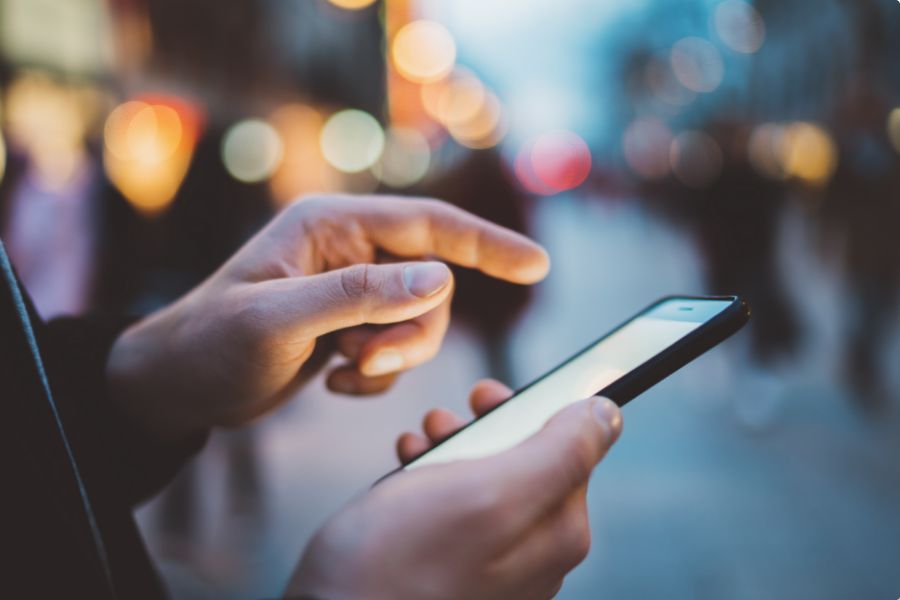 A person using a smartphone on a city street in the evening, with blurred lights and people in the background, creating a bokeh effect.