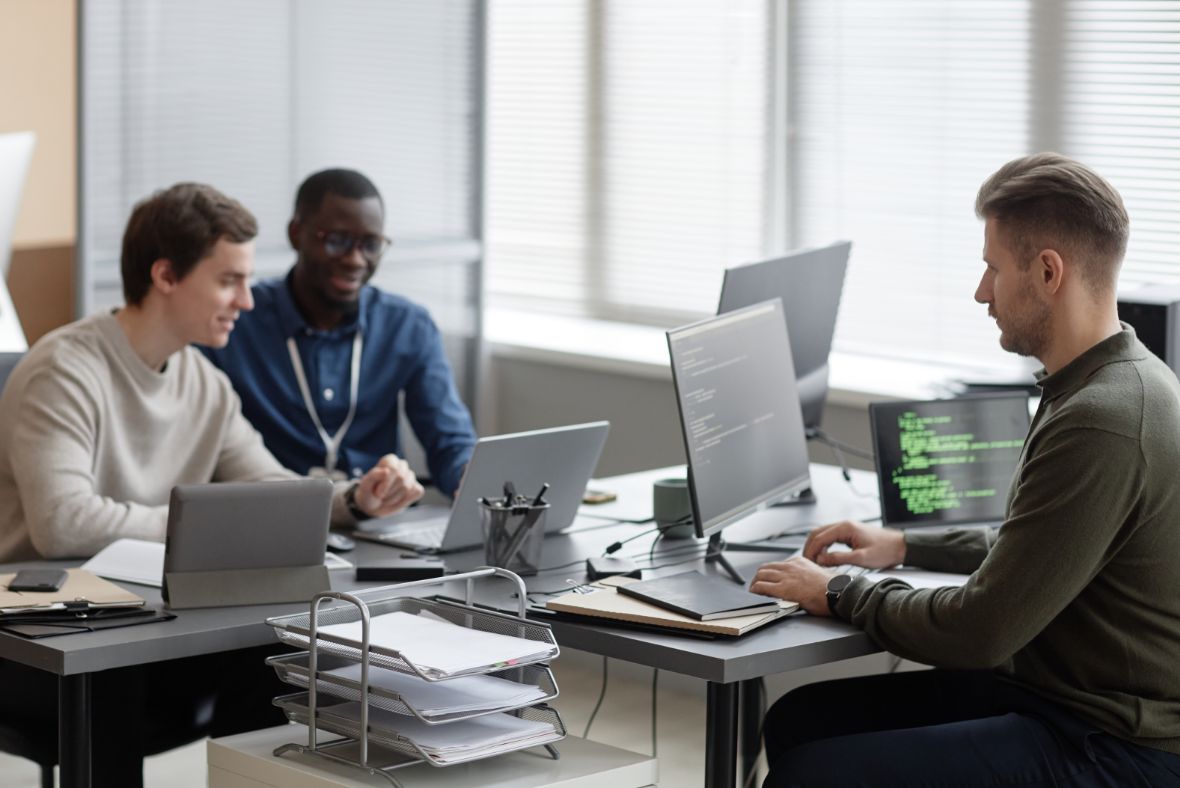 Three men work together in a modern office at desks with laptops and computers. One types code on a monitor while the others talk and smile. Office supplies and paperwork are visible on the desks.