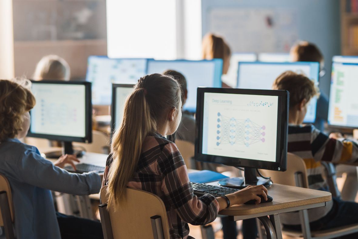 A classroom of students sits at desks, each working on desktop computers. The monitors display diagrams related to deep learning. The scene appears focused and educational, with natural light coming in.