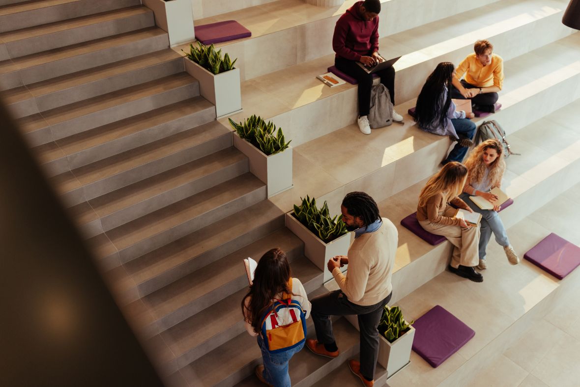 A group of students sit and talk in small groups on tiered steps with potted plants and purple cushions in a bright, modern indoor space. Some read books or use laptops.